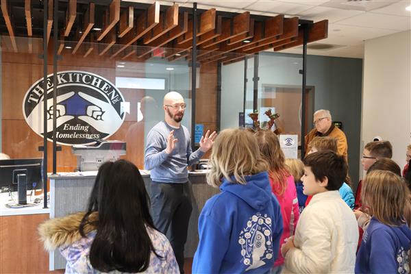  A man stands talking with a crowd of Children. He stands in front of The Kitchen logo.